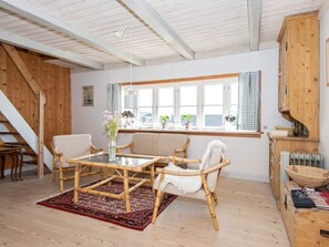 A room with a glass-top table, wicker chairs, a red patterned rug, wooden flooring, a window with curtains, and a staircase.