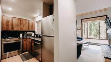 A modern kitchen with stainless steel appliances, wooden cabinets, and a dining area with a view of trees through large windows.