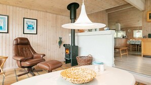 Ett mysigt rum med en fåtölj i läder, ett vitt bord med en korg, en vedeldad kamin och en hängande lampa.