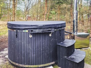 A cylindrical outdoor hot tub with a metal chimney, surrounded by a wooden deck and a forested area.