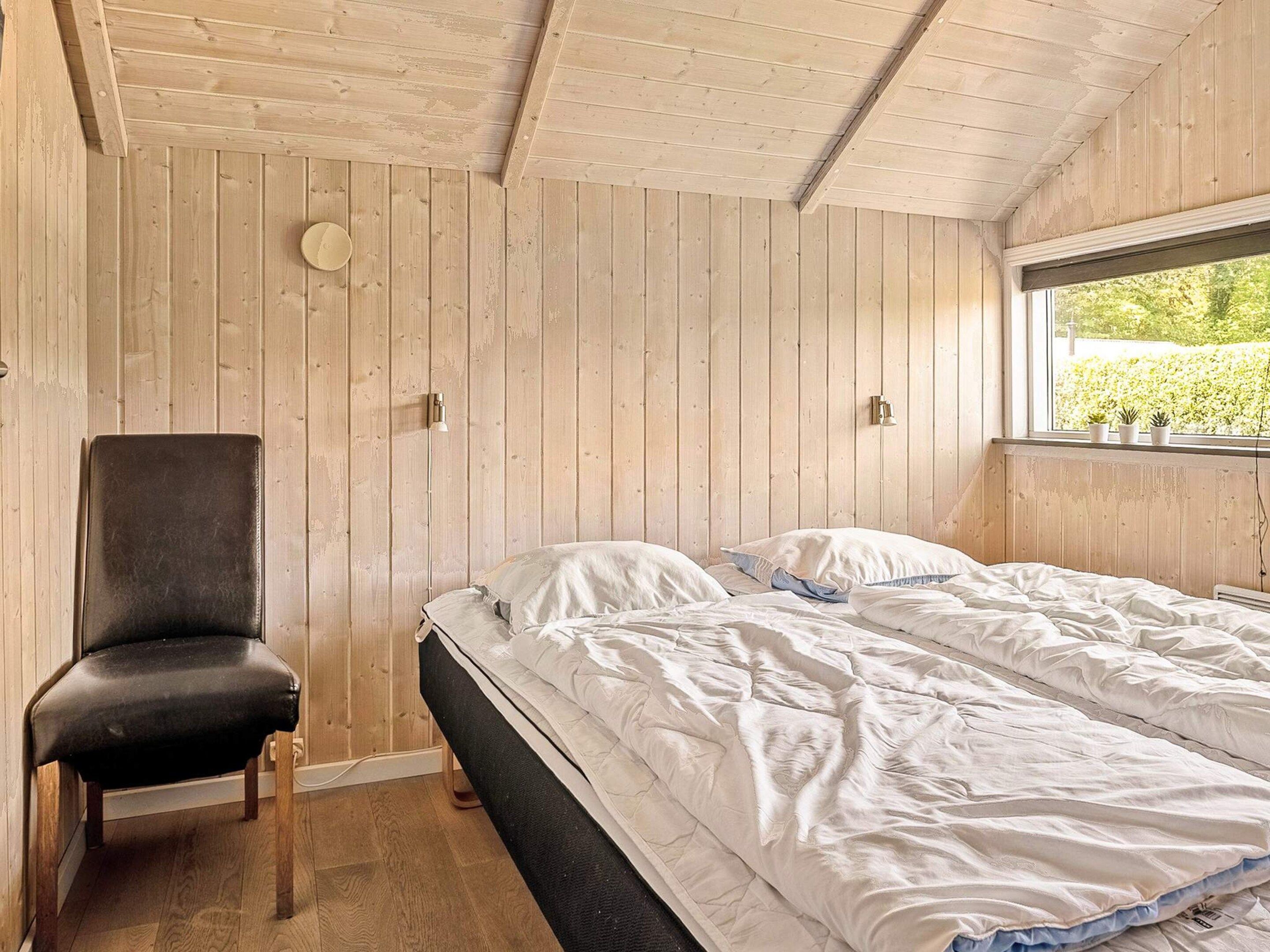 Un dormitorio con techo de madera, una cama con ropa blanca, una silla negra y una ventana con vista a la vegetación.
