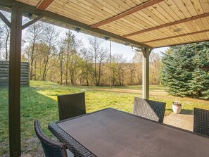 Une terrasse extérieure avec une table et des chaises, entourée d’une pergola en bois, donnant sur une pelouse avec des arbres et un grand conifère.
