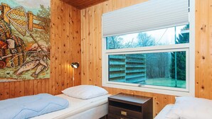 Ein Zimmer mit Holzwänden, zwei Betten, einem Nachttisch, einer Lampe und einem Fenster mit Blick auf Bäume.