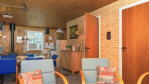 Ein gemütliches Zimmer mit Holzbalkendecke, einer Ziegelwand und einer Holztür. Ein Esstisch mit blauen Stühlen und eine kleine Küchenzeile mit Kühlschrank und Spüle sind vorhanden.