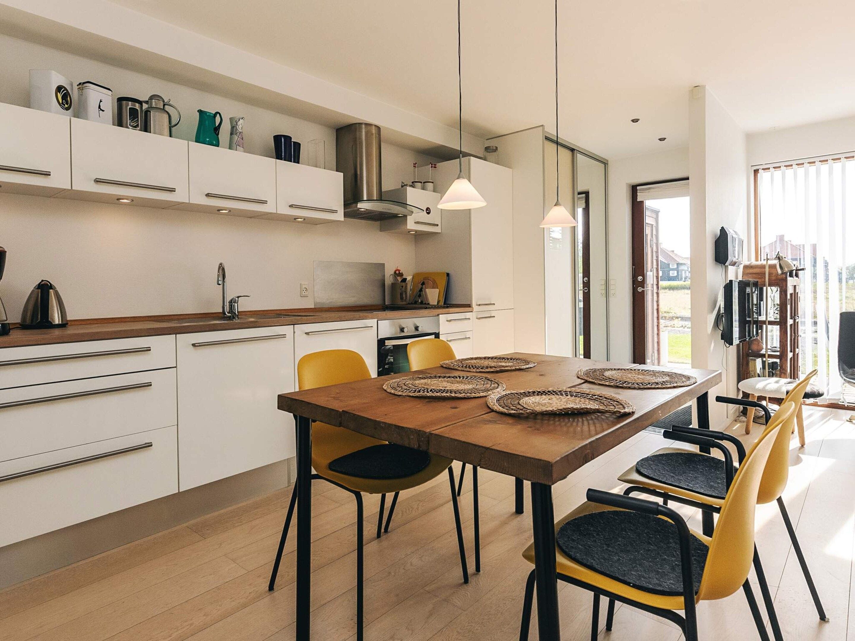 A modern kitchen with white cabinets, a wooden dining table with woven placemats, and a view of the outdoors through a glass door.