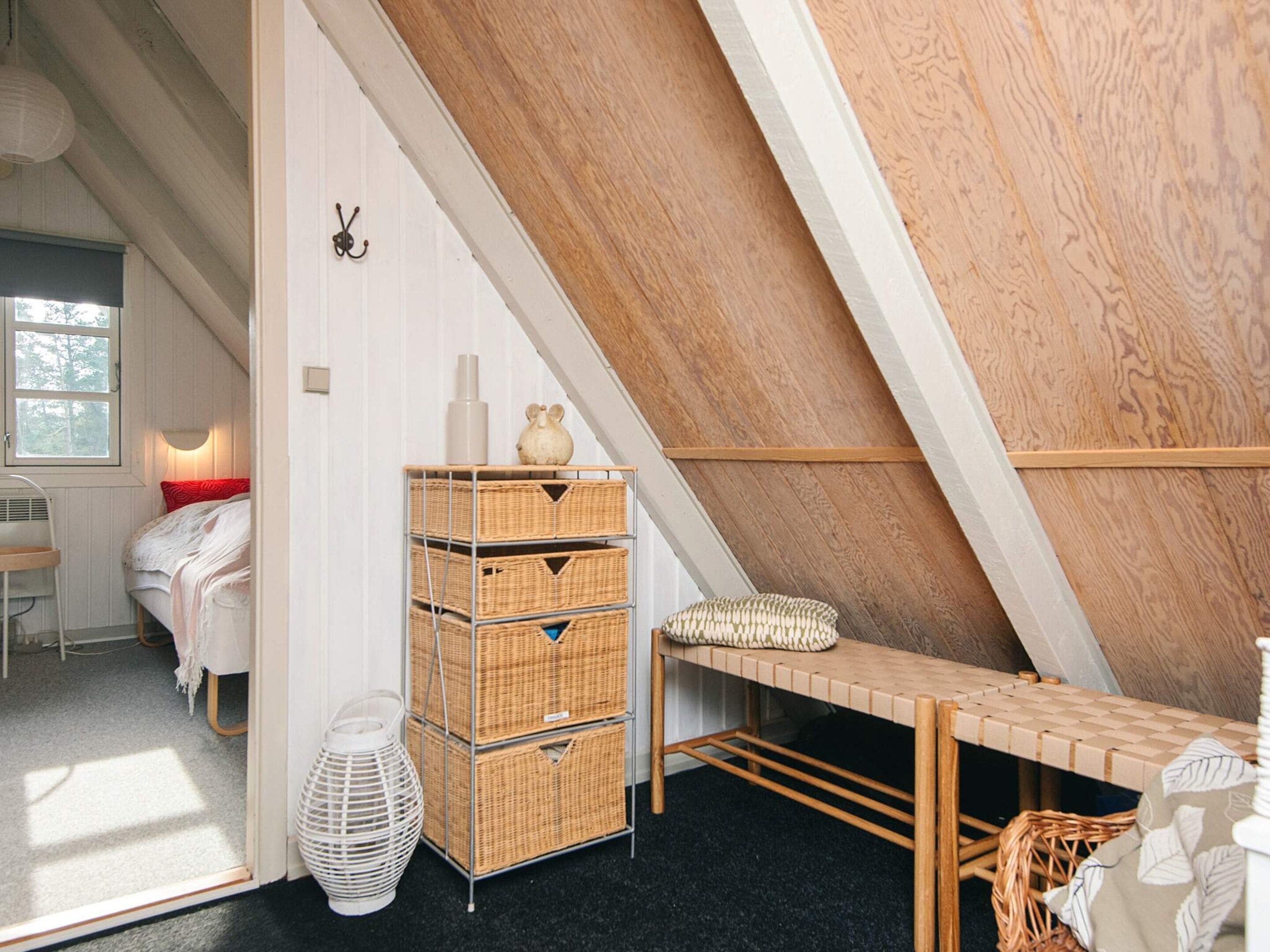 A cozy attic bedroom with a sloped ceiling, a bed with a red pillow, a wooden bench, wicker baskets, and a white lamp.