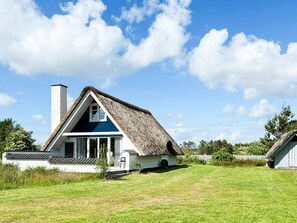 A thatched-roof house with a white exterior and a blue window, surrounded by a grassy yard and a white fence.