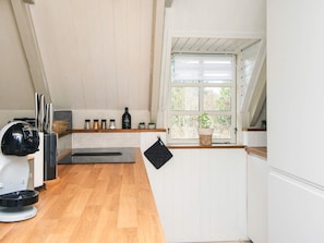 A kitchen with a wooden countertop, a coffee machine, a window with a view of trees, and a shelf with kitchen utensils.