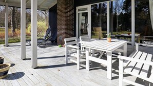 Un patio al aire libre con una mesa y sillas blancas, una chimenea de ladrillo y un ventanal de vidrio que da a un jardín.