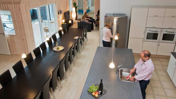 Een moderne keuken met een lang eiland, een zithoek met een donker houten tafel en stoelen, en uitzicht op de omgeving door grote ramen.