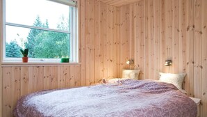Een slaapkamer met houten paneelwanden, een bed met een geborduurde spreidekker, twee bedlampen en een raam met uitzicht op groen.