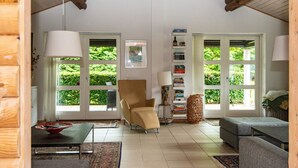 Un salon spacieux avec un plafond haut, des poutres en bois, un canapé confortable, un fauteuil, une table basse, une bibliothèque et de grandes fenêtres donnant sur un paysage verdoyant.