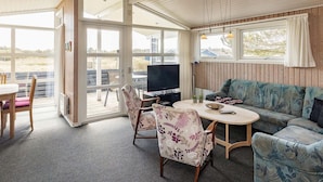 Ein gemütliches Zimmer mit Esstisch, Stühlen, einer Couch und einem Fernseher. Große Fenster bieten einen Ausblick ins Freie.