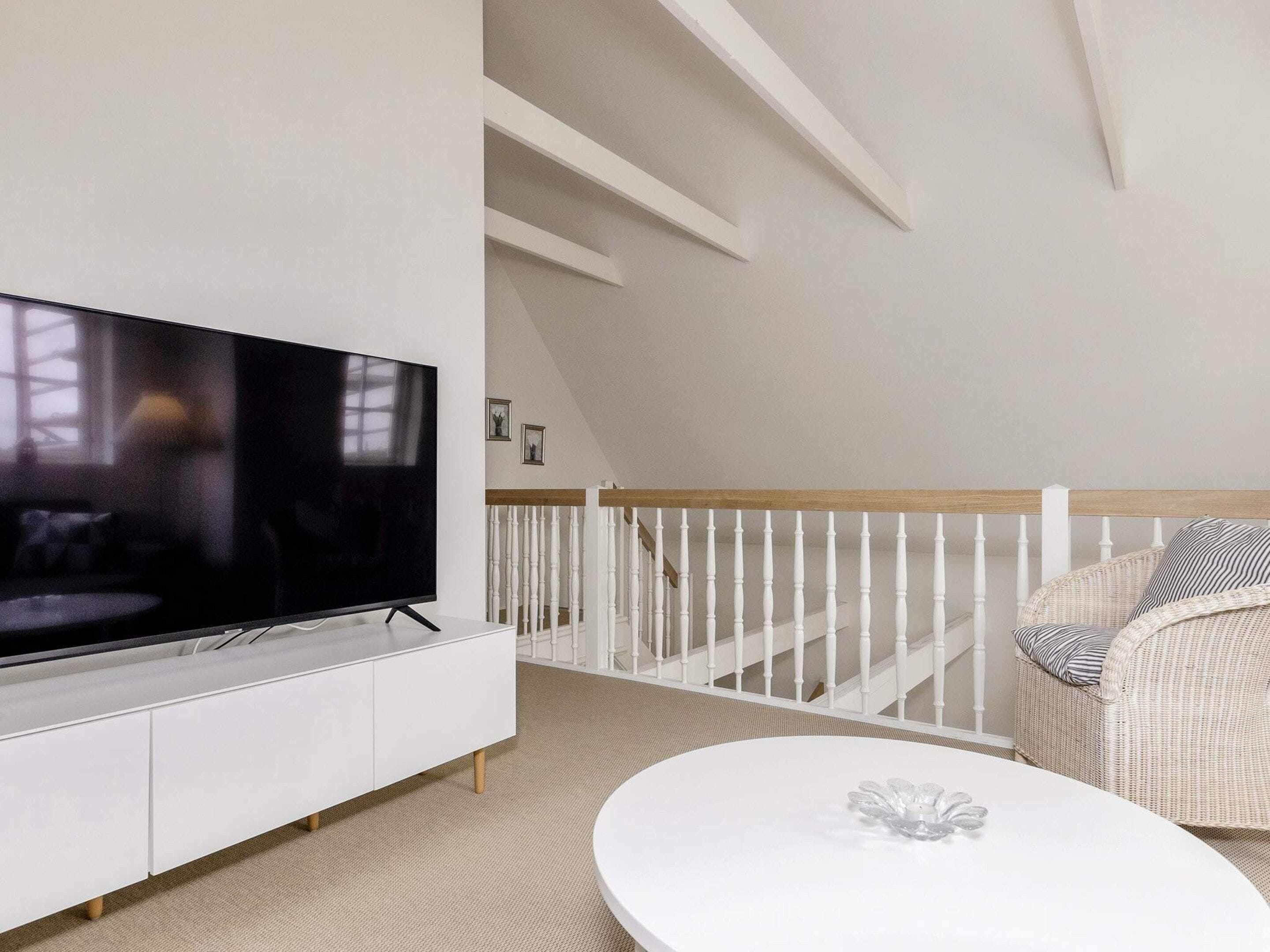 Un salon moderne avec un téléviseur à écran plat posé sur une console blanche, une table basse ronde ornée d’un objet décoratif en verre, un fauteuil en osier avec un coussin à rayures et une rambarde blanche menant à une mezzanine.
