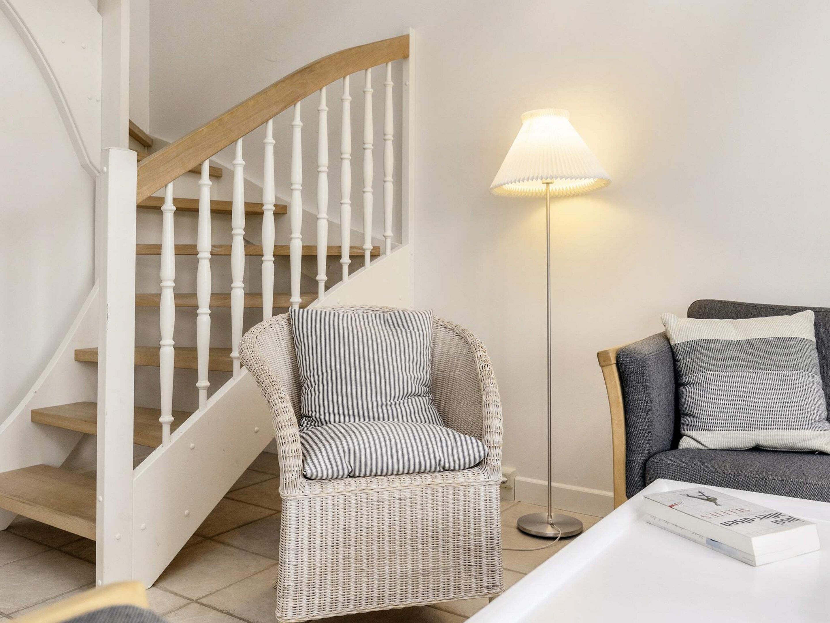 A wicker chair with striped cushions, a grey sofa with patterned cushions, a white table with a book, and a wooden staircase with white balusters.