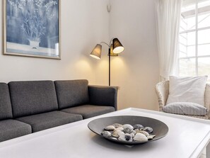 Una sala de estar moderna con un sofá gris, una mesa de centro blanca con conchas marinas, una lámpara de pie y una silla de mimbre con cojines.