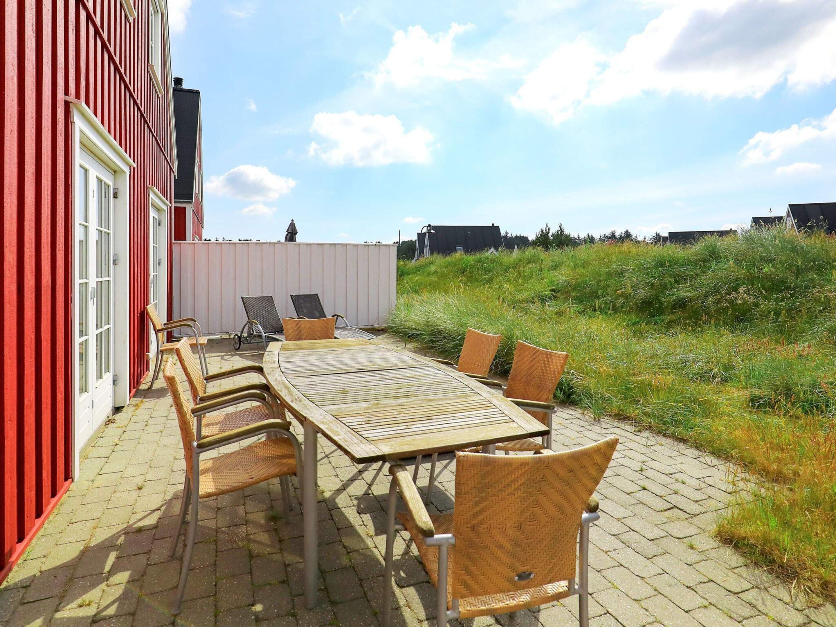 Eine Terrasse im Freien mit einem Holztisch und Korbstühlen, neben einem roten Gebäude mit weißen Verzierungen.
