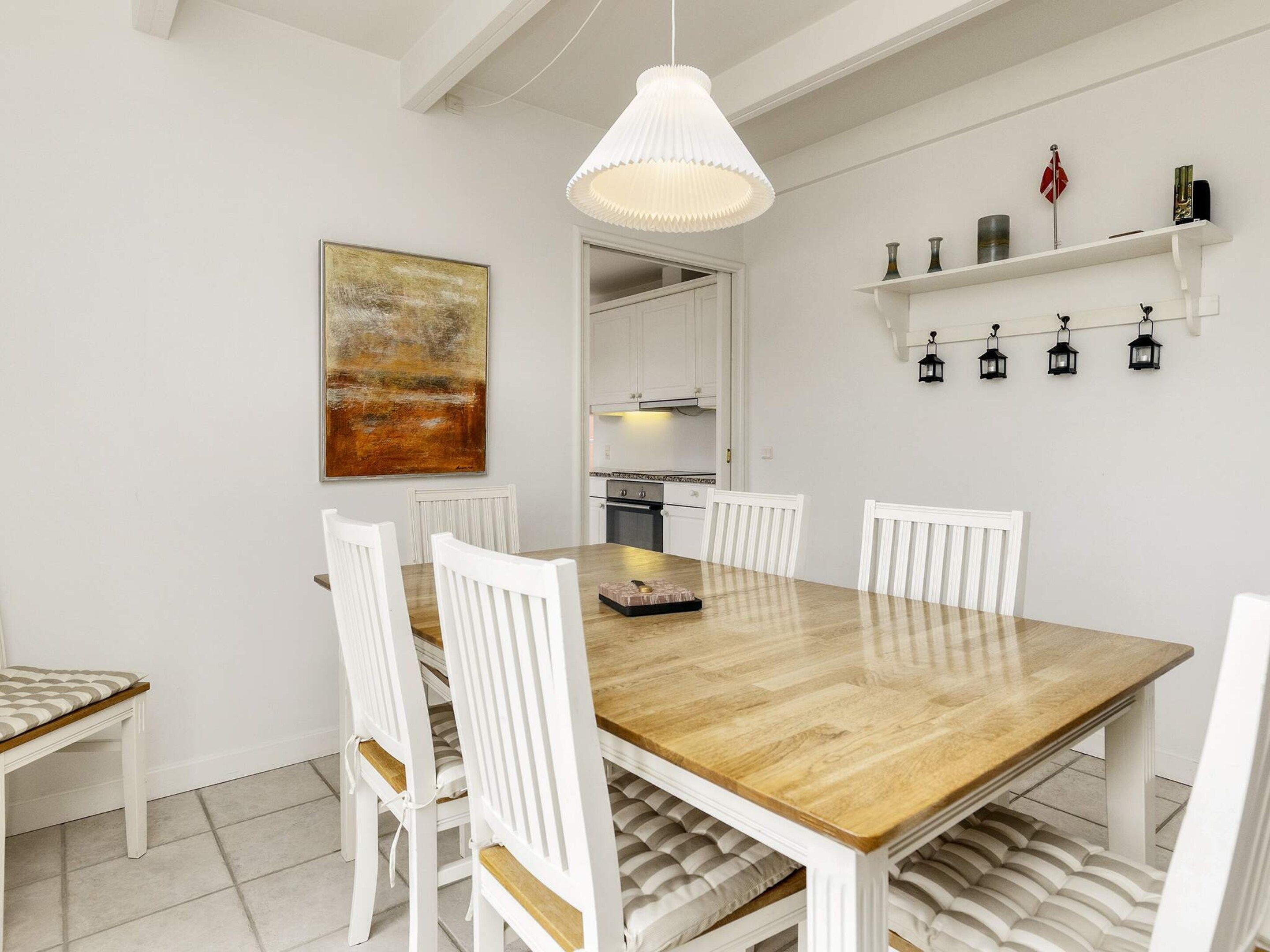 A dining area with a wooden table and white chairs, a pendant light, a painting, and a shelf with decorative items.