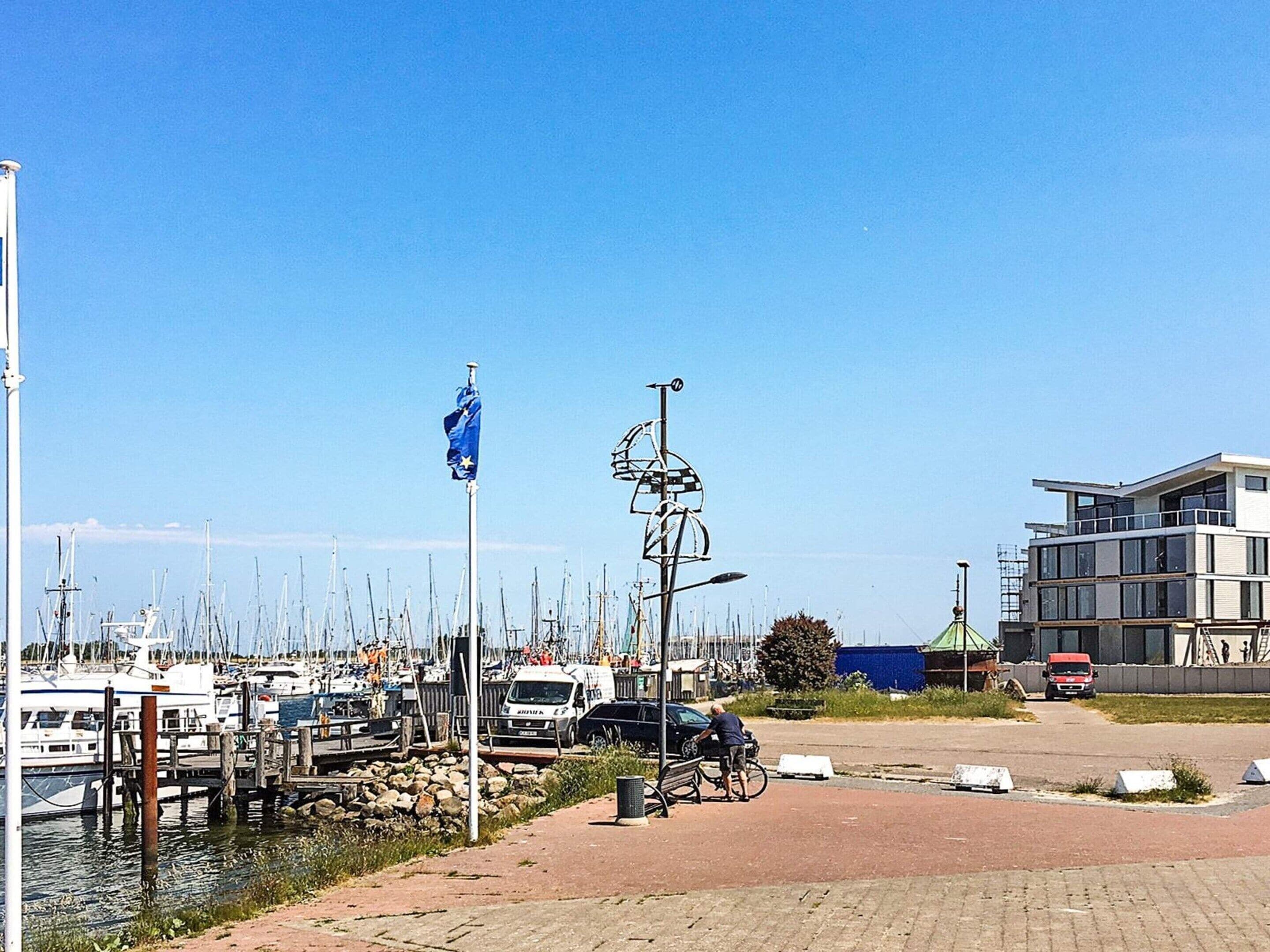 A marina with boats docked, a flagpole with a flag, a bicycle, and a modern building in the background.