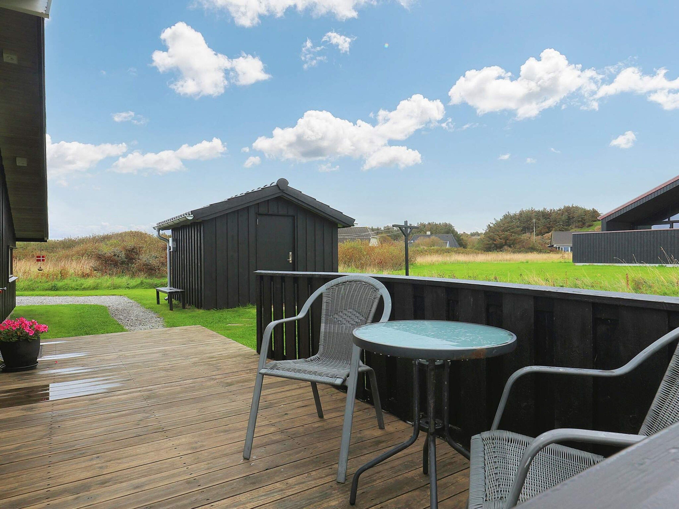 En træterrasse med udendørs møbler, et lille skur og en græsplæne i baggrunden.