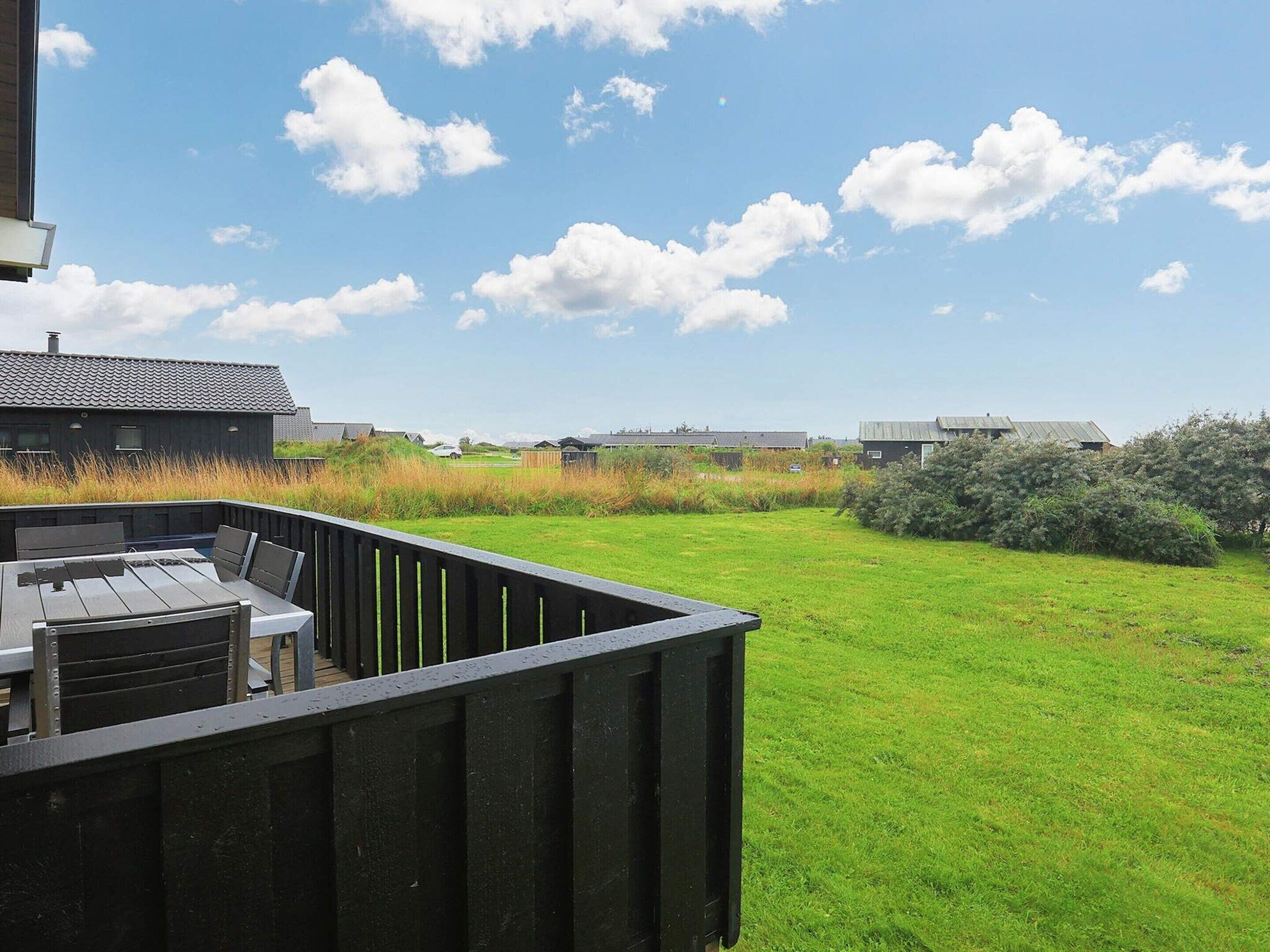 A patio area with a table and chairs, surrounded by a grassy lawn and a view of a row of houses under a clear sky.
