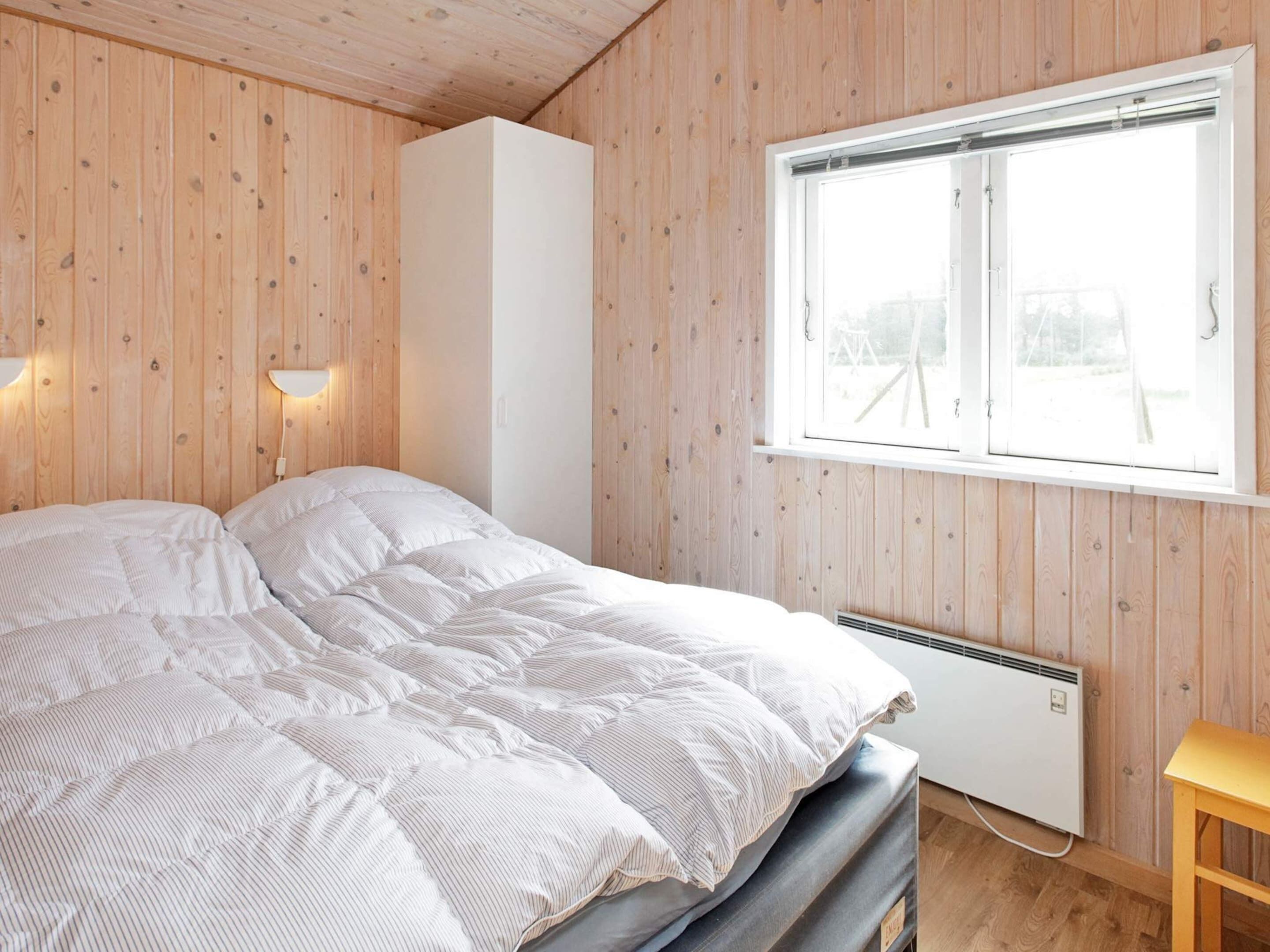 A bedroom with a bed, a window, a wall-mounted heater, and a wooden bench.