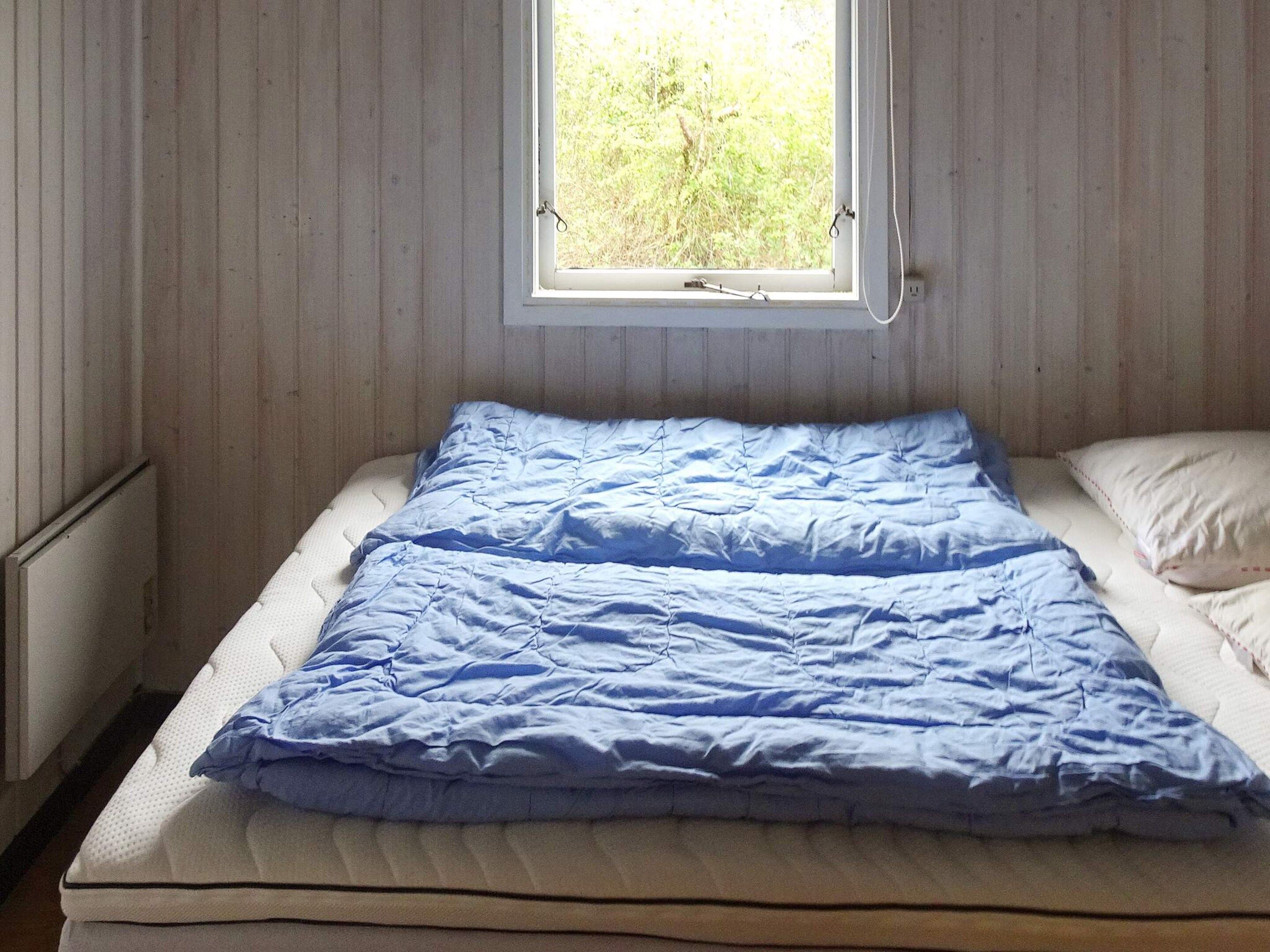 A single bed with a blue blanket, a white pillow, and a window with a view of greenery outside.