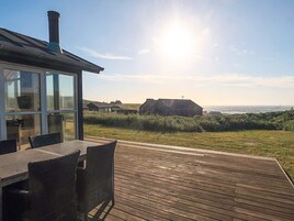 Una terraza de madera con mobiliario exterior, una sala con paredes de vidrio y vistas al océano y edificios distantes.