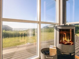 Una estufa de leña con leños ardiendo en su interior, una olla con tapa y una ventana con vistas a un área cubierta de césped.