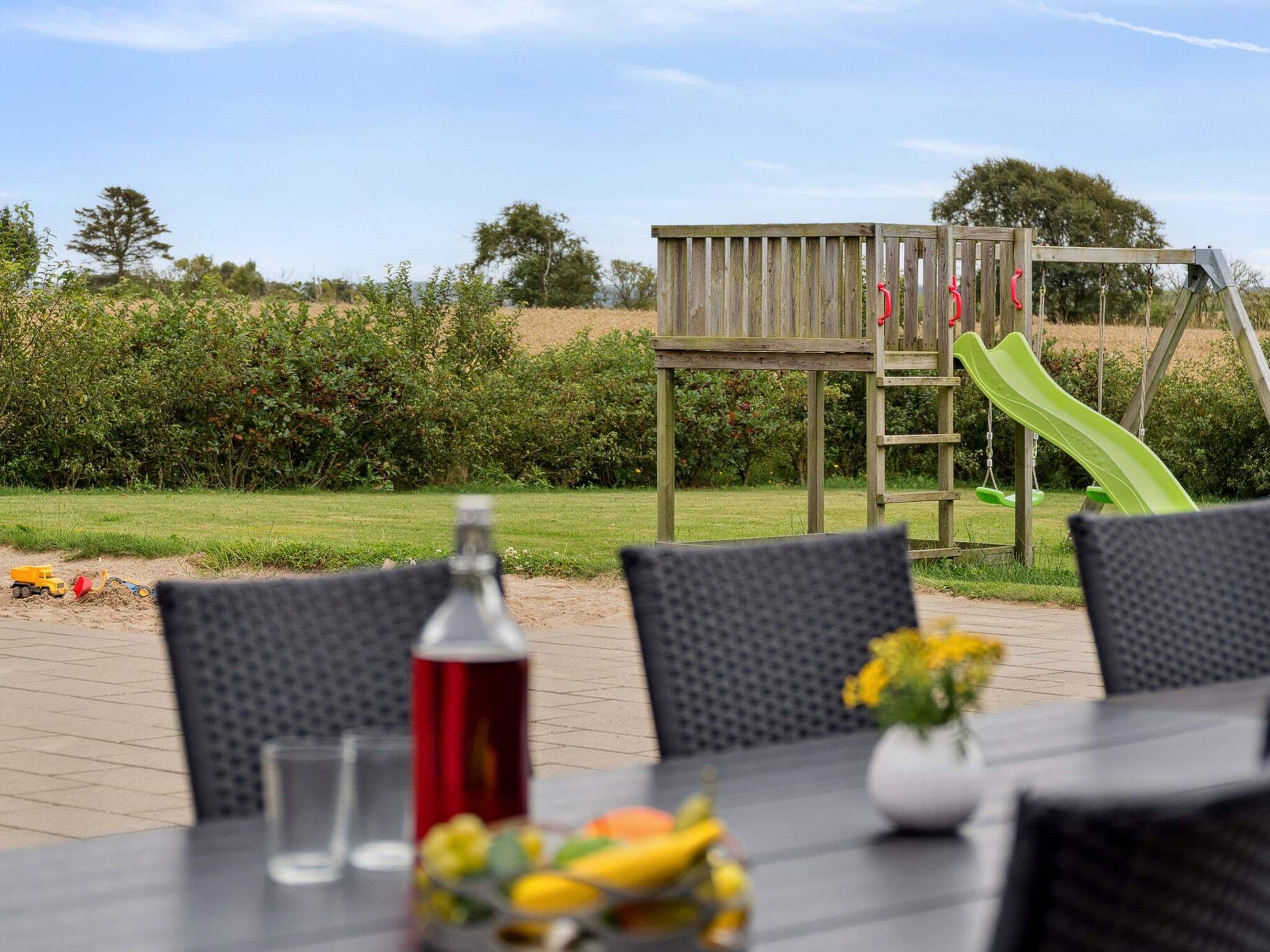 An outdoor dining area with a table set for two, a bottle, and a fruit bowl. In the background, there is a wooden play structure with a slide.