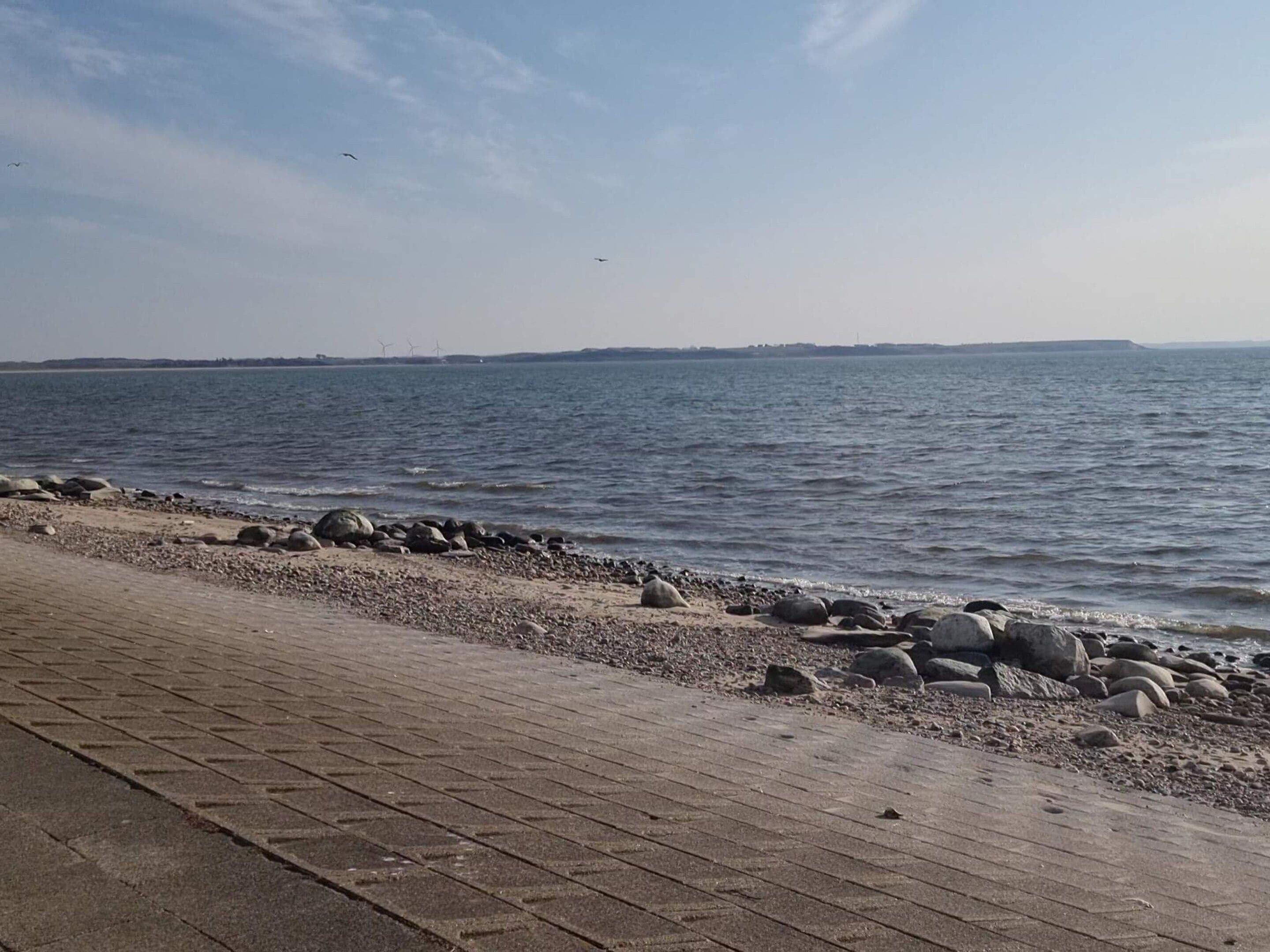 A paved walkway beside a body of water with rocks and distant wind turbines.