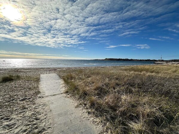 Un sentier côtier qui mène à une plage, avec un ciel dégagé et une vue sur la terre ferme au loin.