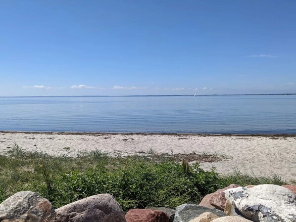 Ein Strand mit ruhigem Wasser, einem entfernten Segelboot und einem felsigen Vordergrund.