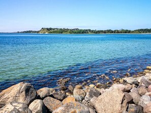 A rocky shoreline with clear water and a distant tree-covered landmass. - 5 Person Holiday Home in Jaegerspris (Jægerspris)