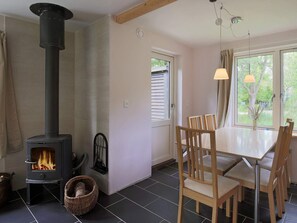 A room with a wood-burning stove, a dining table with chairs, a wicker basket, and a window with curtains.