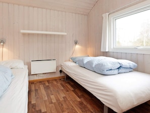 A room with two beds, a window, a wall-mounted heater, and a wooden floor.