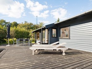 A wooden deck with a lounge chair, a table, and chairs, and a building with large windows and a covered porch.