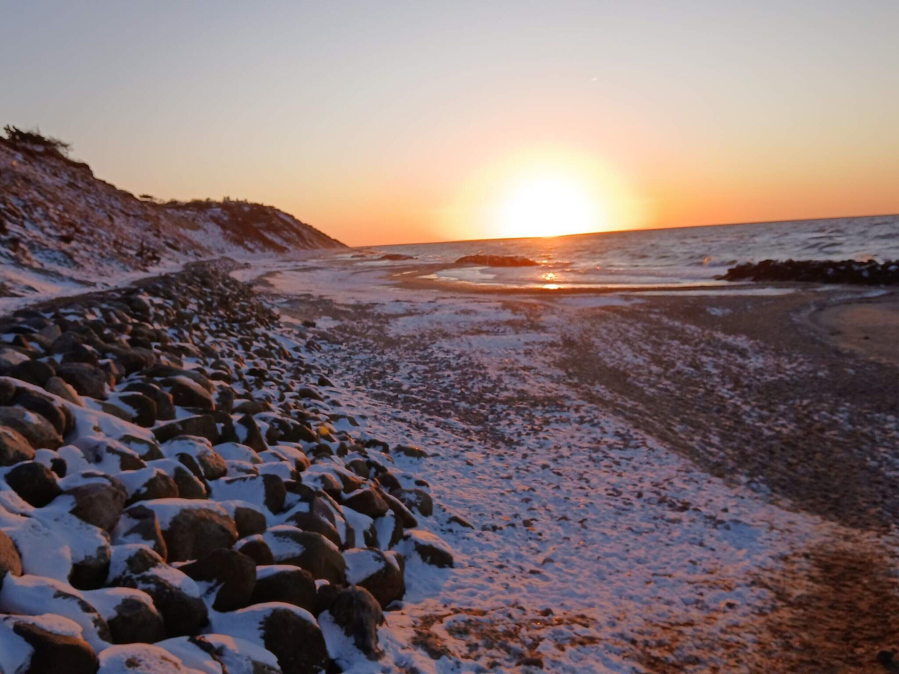 En stenet kystlinje med snedækkede sten og en solnedgang over havet.