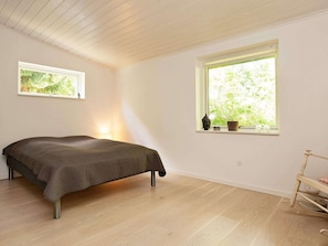 A bedroom with a bed, a window with a view of greenery, a small shelf with items, and a wooden floor.
