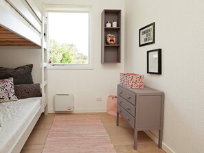 Quarto com uma cama de solteiro e beliche, cômoda, radiador, janela com vista e prateleira com objetos decorativos.
