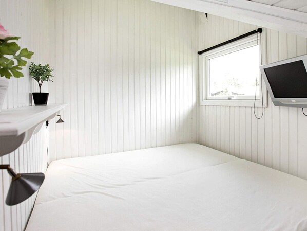 A small, white-walled room with a single bed, a mounted TV, a window with a blind, and a small shelf with a plant.