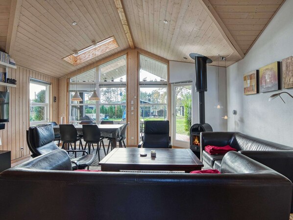 A cozy living room with a fireplace, wooden ceiling, and large windows offering a view of the outdoors.