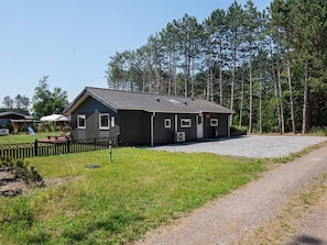 A small, single-story building with a gravel driveway, surrounded by a grassy area and tall trees.