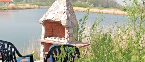 An outdoor stone barbecue grill with a chimney, a chair, and a view of a lake.