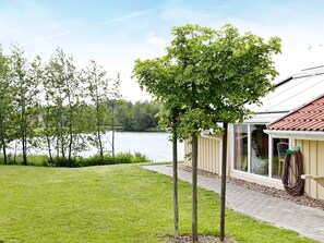 A house with a glass door, a garden with grass and trees, and a lake in the background. - Holiday Home in Otterndorf (Otterndorf)