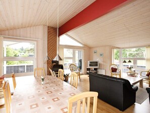 A spacious living room with a large wooden table, comfortable seating, a fireplace, and large windows offering a view of greenery.