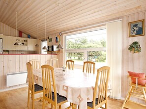 A dining area with a round table and chairs, a kitchen with wooden cabinets, and a large window with a view of greenery.