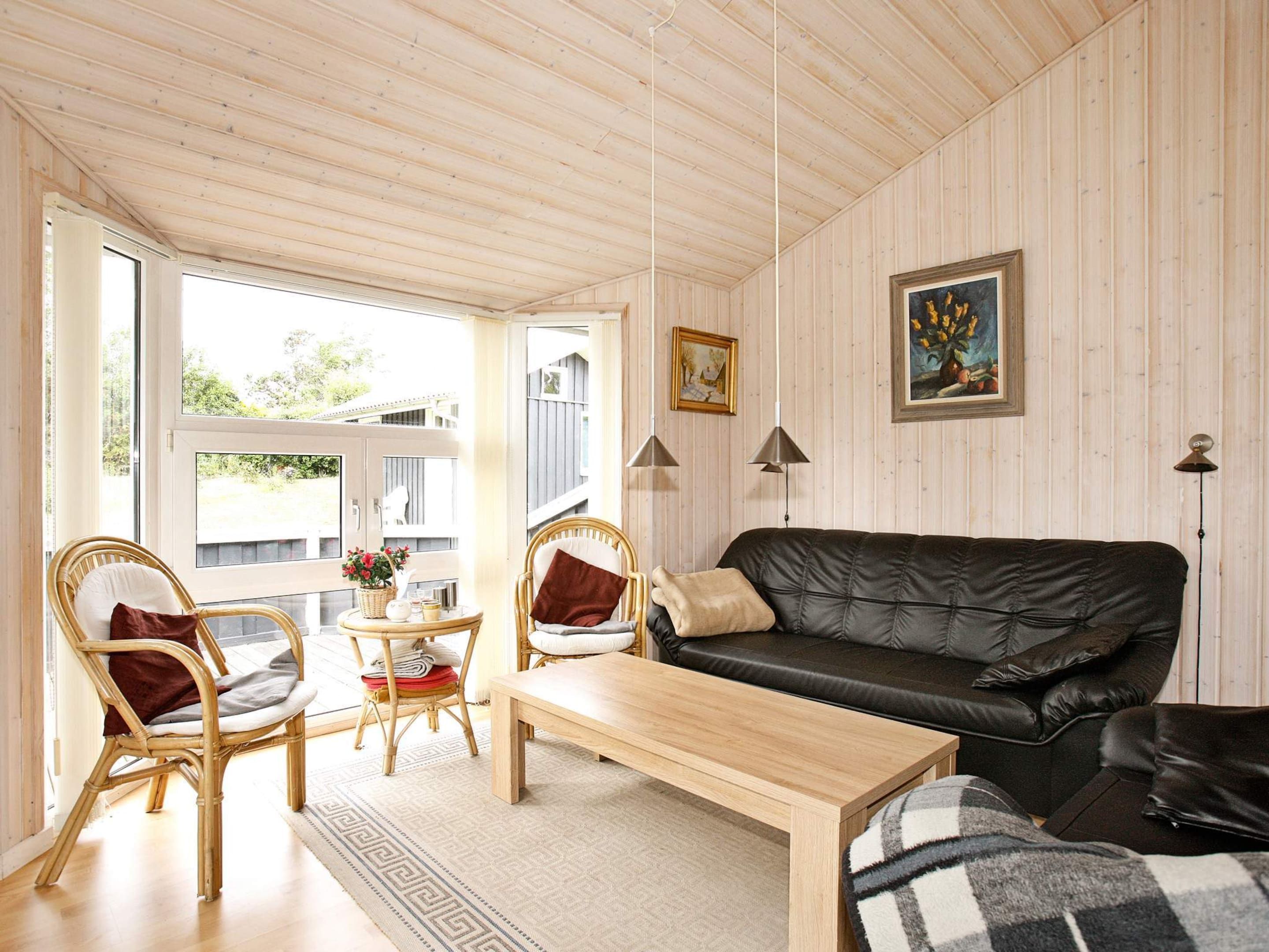 A cozy living room with a wooden ceiling, a black leather sofa, two wicker chairs, a wooden coffee table, and a view through a large window.