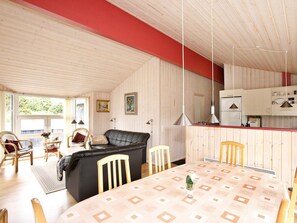 A cozy living room with a black leather sofa, wooden chairs, a dining table with a patterned tablecloth, and a kitchen area with white cabinets and hanging pendant lights.