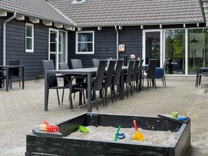 Un espace extérieur aménagé en patio, avec une longue table et des chaises, un bac à sable rempli de jouets, et un bâtiment doté de fenêtres et d’une porte.